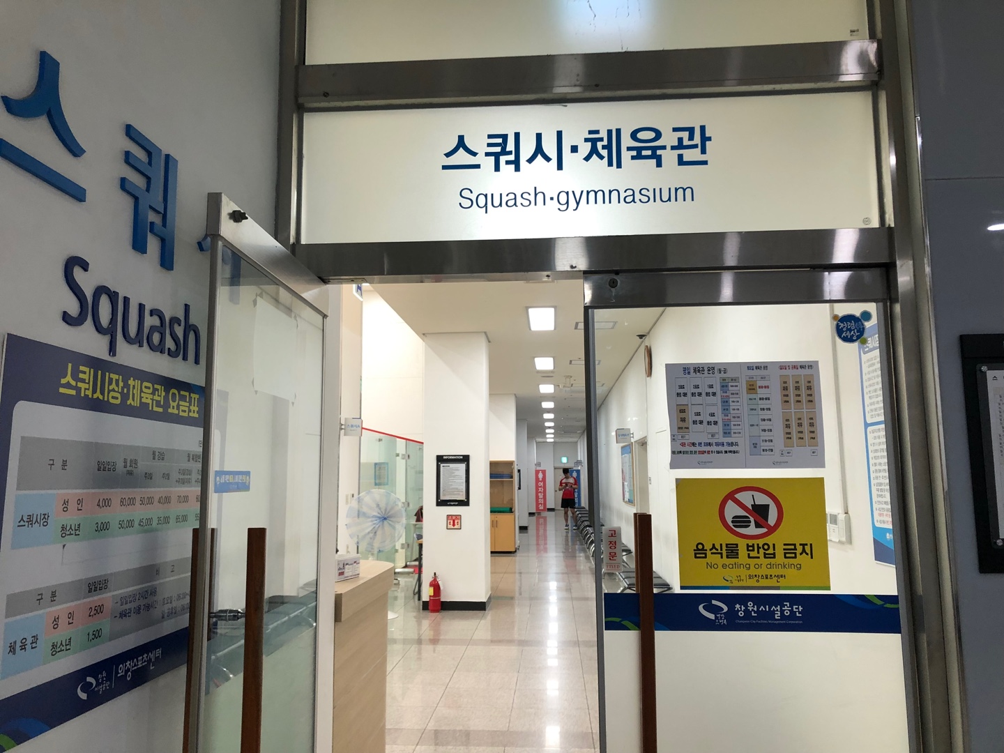 스쿼시 및 체육관 입구를 담은 사진이며, 입구 좌측에는 스쿼시장과 체육관 요금표가, 입구 문에는 음식물 반입금지 안내가 표시되어 있고 우측에는 프로그램 안내판이 비치되어 있습니다.