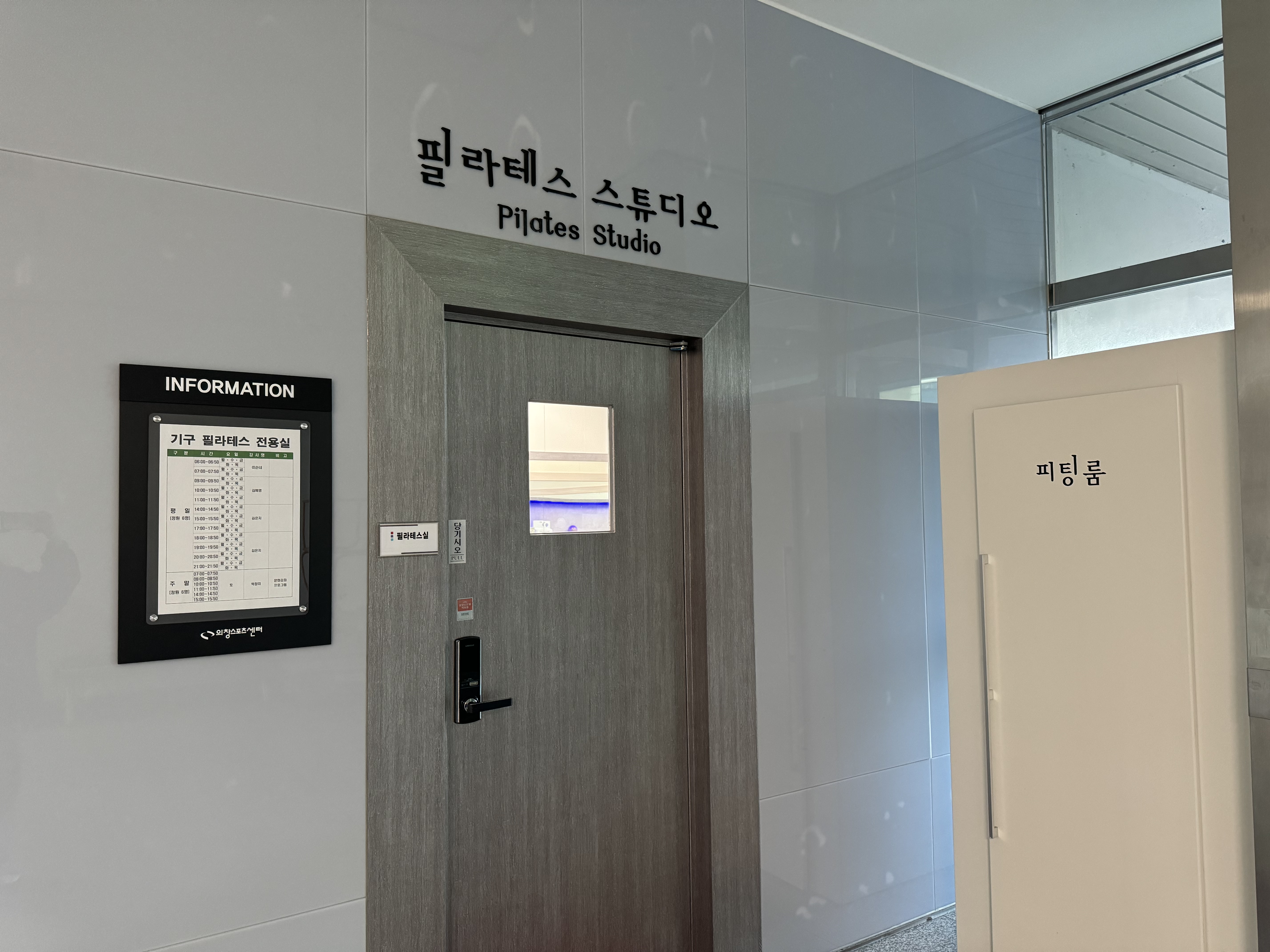 필라테스 스튜디오의 입구 사진이며, 입구를 기준으로 좌측에 월별 프로그램 관련 안내판이 비치되어 있습니다. 또한 입구 우측에는 피팅룸(탈의실)이 있습니다.