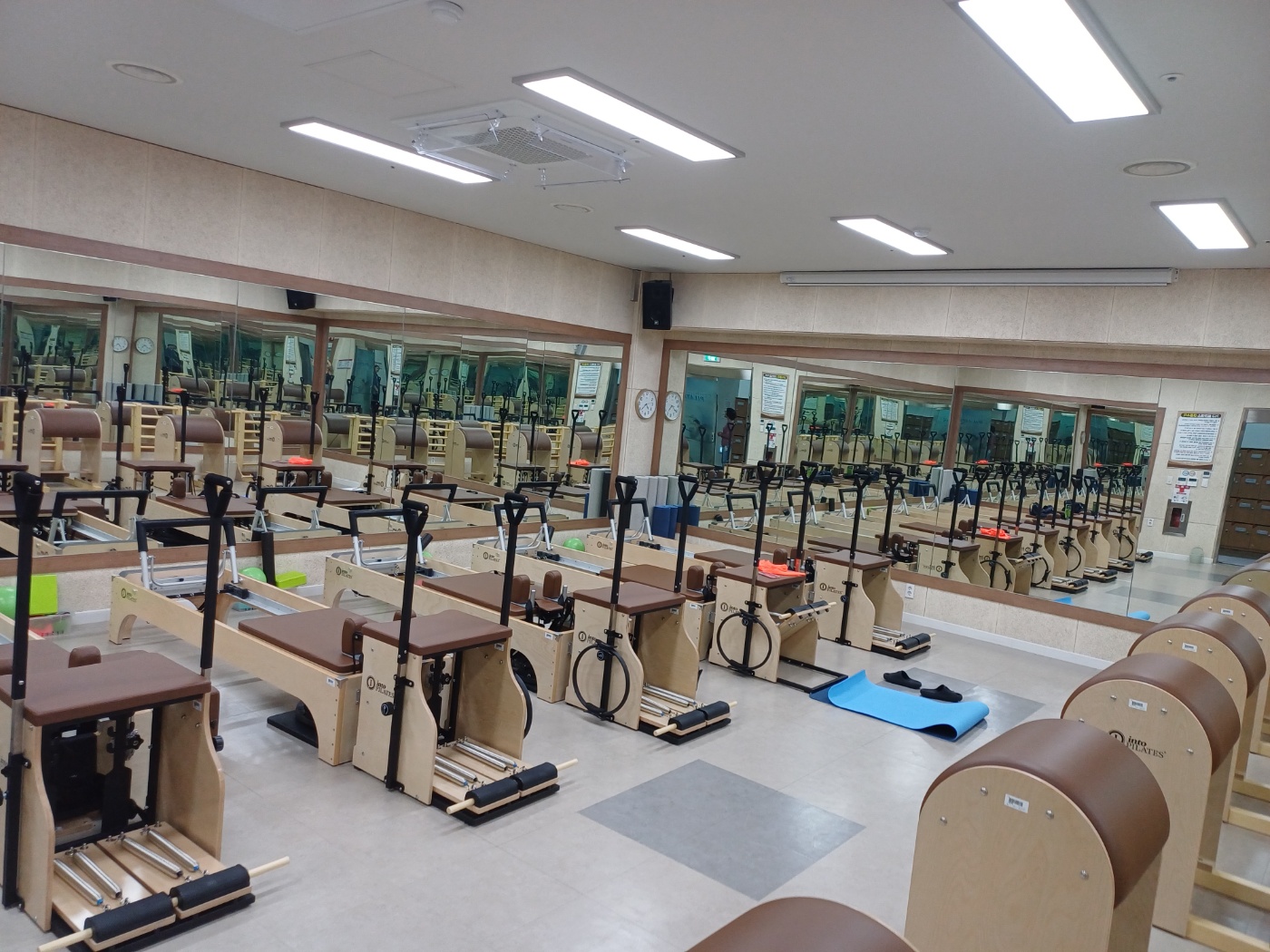 필라테스 스튜디오 안쪽 가장자리를 방향으로 촬영한 내부 사진이며, 안쪽 가장자리를 기준으로 좌우측 벽면에 거울이 설치되어 있고, 중앙에는 기구들이 나열되어 있습니다.