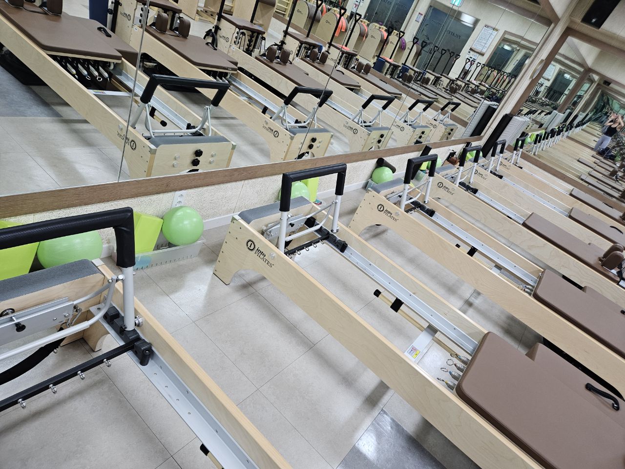 필라테스 스튜디오 내부 기구(리포머) 쪽에서 안쪽 대각선 방향으로 촬영한 사진이며, 벽면 거울 앞 기구가 일렬로 나열되어 있습니다.